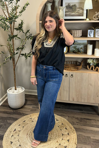 Woman in black top and blue jeans standing in a room with a plant and shelves.