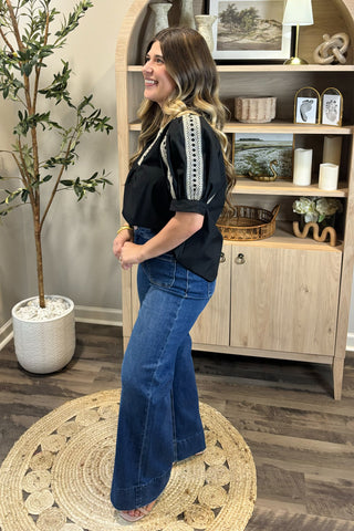 Woman in black top and blue jeans standing in a room with wooden flooring and a plant.