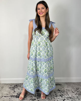 Smiling woman standing against a neutral wall wearing a flowy tiered maxi dress with a fresh green-and-blue floral print, featuring sweet striped bow-tie shoulder straps, a square neckline, and contrasting striped banded tiers for a charming, cottage-inspired look, styled with nude block-heel sandals and delicate gold jewelry for an effortless spring-to-summer outfit.