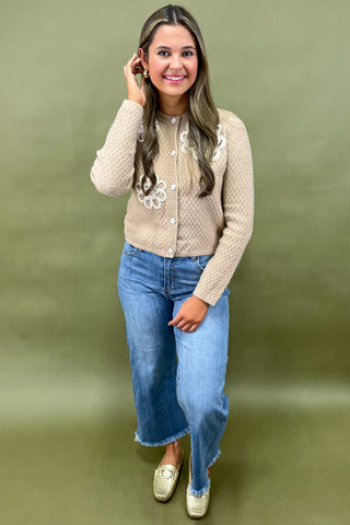 Woman wearing a beige knitted cardigan with floral patterns and blue jeans against a green background