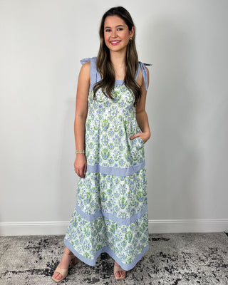 Smiling woman standing against a neutral wall wearing a flowy tiered maxi dress with a fresh green-and-blue floral print, featuring sweet striped bow-tie shoulder straps, a square neckline, and contrasting striped banded tiers for a charming, cottage-inspired look, styled with nude block-heel sandals and delicate gold jewelry for an effortless spring-to-summer outfit.