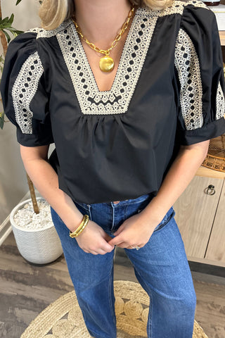 Person wearing a black blouse with white lace details and blue jeans indoors.