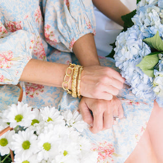 Close-up of hands with gold bracelets and a ring, surrounded by flowers.