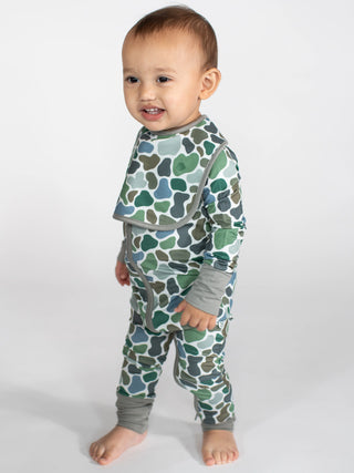 A baby wearing a long-sleeve camouflage print pajama set with a bib, standing against a light gray studio background.