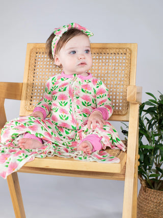 A baby wearing a pink and green floral print footless one-piece pajama and matching headband, sitting on a wooden cane-back chair.