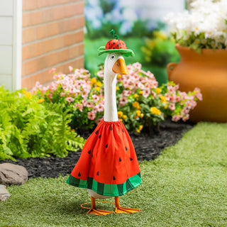 Decorative goose in a watermelon costume standing on grass with flowers and plants in the background.