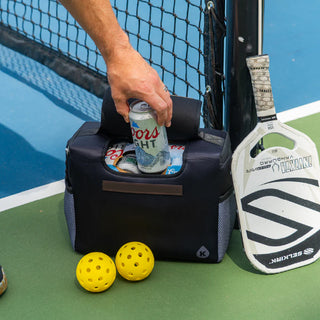 Person opening a cooler on a tennis court with a can of beer, pickleball paddles, and balls.
