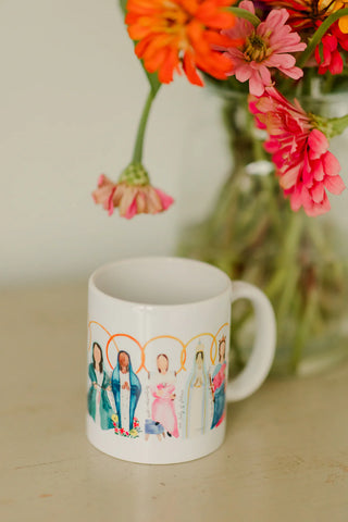 White mug with a colorful illustration of people on a beige surface with flowers in the background