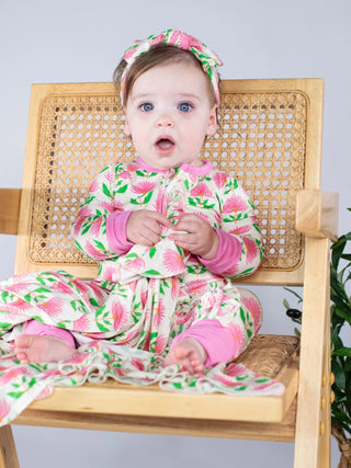 A baby wearing a pink and green tropical print one-piece pajama set with a matching headband, sitting on a wooden cane chair.