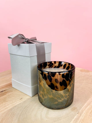 Candle with leopard print design next to a gift box on a wooden surface with a pink background