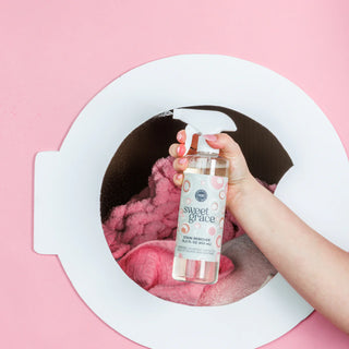 Hand holding a bottle labeled 'sweet grace' over a washing machine with pink fabric inside on a pink background.