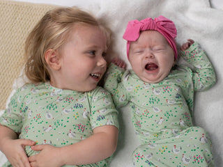 Two children in matching green outfits with a pink headband, lying on a white blanket.