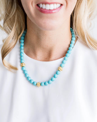 Person wearing a turquoise beaded necklace with gold accents on a white background