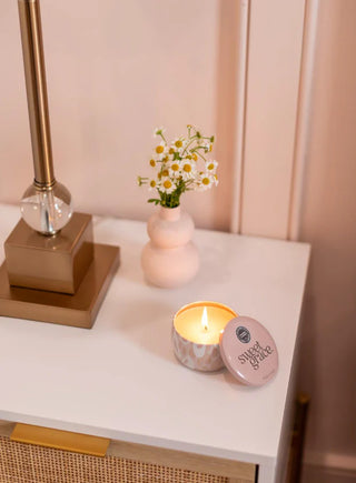 Candle with a decorative lid on a table next to a small vase with flowers, against a light pink wall.