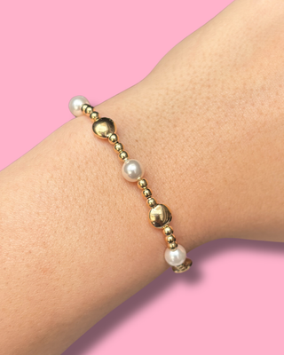 Bracelet with gold beads and pearls on a wrist against a pink background