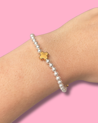 Pearl bracelet with a gold cross charm on a wrist against a pink background