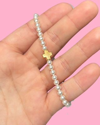 Pearl bracelet with a gold cross held in a hand against a pink background