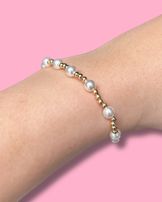 Pearl and gold beaded bracelet on a wrist against a pink background