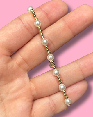 Hand holding a bracelet with alternating gold and pearl beads against a pink background