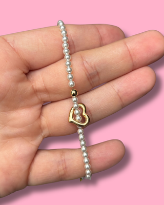 Pearl bracelet with a gold heart charm held in a hand against a pink background