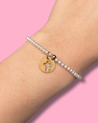 Pearl bracelet with a gold disc charm featuring a cross on a pink background