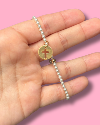 Pearl bracelet with a gold tag featuring a cross on a hand against a pink background