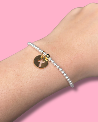 Pearl bracelet with a gold disc featuring a cross on a wrist against a pink background