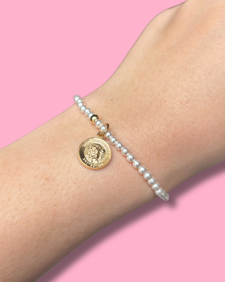 Pearl bracelet with a gold coin charm on a wrist against a pink background
