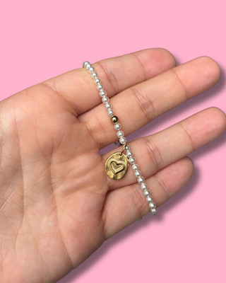 Hand holding a pearl bracelet with a gold heart charm against a pink background