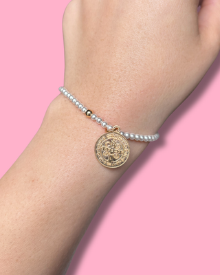 Gold coin charm on a pearl bracelet against a pink background