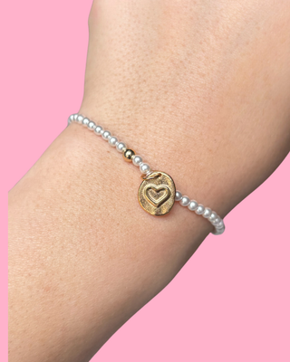 Gold heart charm bracelet on a wrist against a pink background