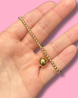 Gold beaded necklace held in a hand against a pink background