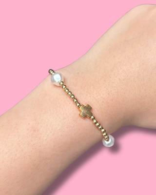 Bracelet with gold beads and a cross on a wrist against a pink background