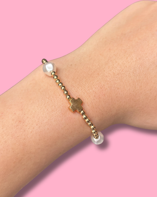 Bracelet with gold beads and a cross on a wrist against a pink background