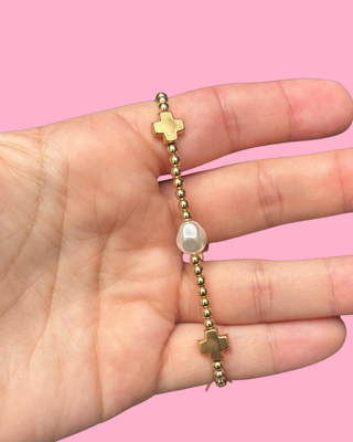 Gold bracelet with crosses and a pearl held in a hand against a pink background