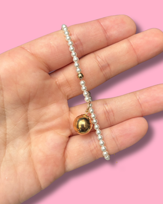 Hand holding a pearl bracelet with a gold bead against a pink background