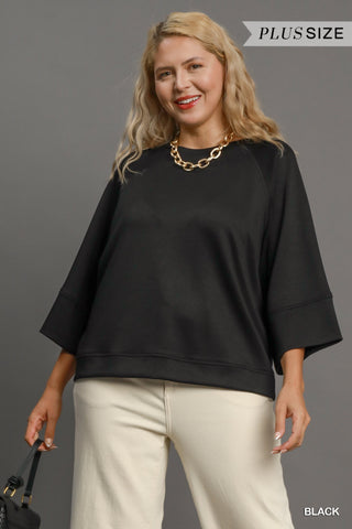 Woman wearing a black plus-size top and beige pants against a gray background