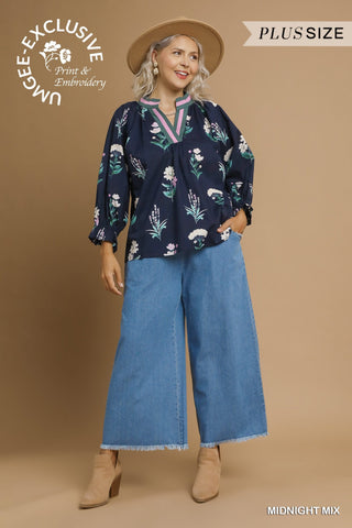 Woman wearing a floral blouse and blue jeans with a brown hat on a beige background, featuring Umgee branding.
