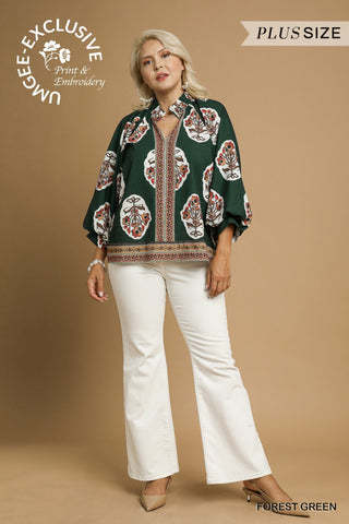 Woman wearing a forest green patterned blouse with white pants on a beige background, featuring Umgee Exclusive branding.