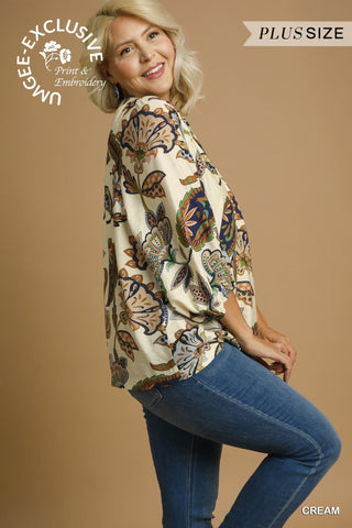 Woman wearing a plus-size cream blouse with floral patterns and blue jeans on a beige background.