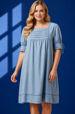 A woman modeling a light blue chambray dress with a square neckline, gathered chest detailing, short puff sleeves with tie cuffs, and a knee-length skirt, set against a dark blue background with window shadow overlays.
