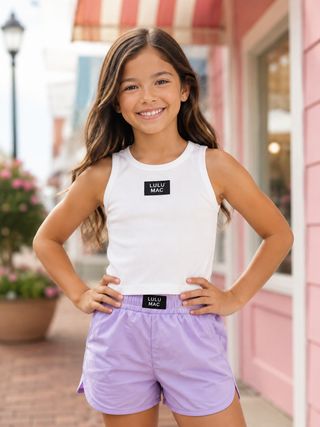 wearing a white sleeveless tank top with a black Lulu Mac logo patch on the chest and lavender athletic shorts wit
