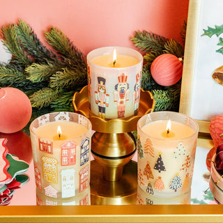 Three lit candles with festive designs on a gold stand, surrounded by Christmas decorations.