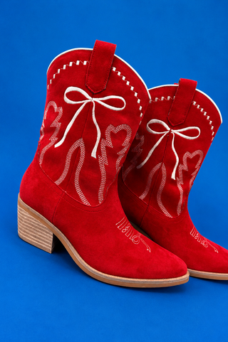 A pair of red suede cowboy boots with white embroidered western-style patterns and bows on the shafts, shown against a sol
