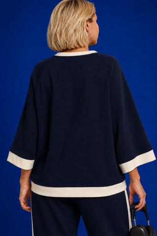 A woman shown from the back wearing a navy blue knit top with contrasting cream trim at the collar, against a solid blue studio background.