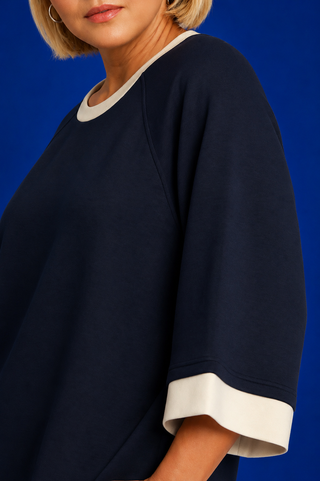 A close-up of a person wearing a navy blue oversized top with contrasting white ribbed trim at the crew neck and sleeve cuffs, set against a solid blue studio background.