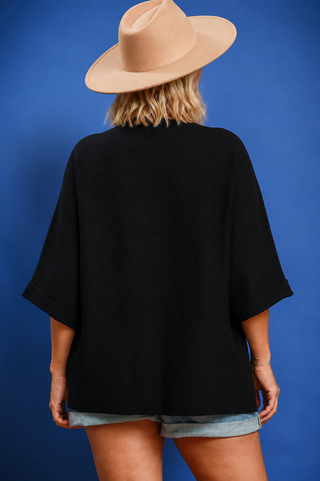 A woman shown from behind wearing a loose black short-sleeve top, denim shorts, and a beige wide-brim hat against a solid blue background.