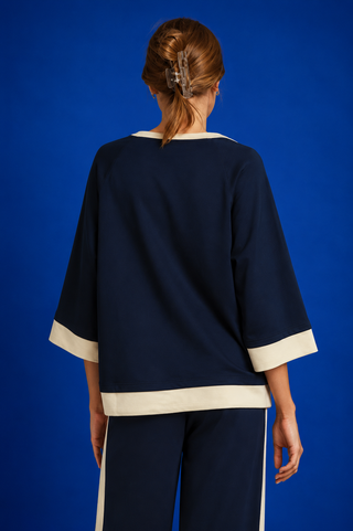 A woman shown from the back wearing a navy blue top with cream trim and matching navy pants with cream side stripes, set against a solid blue studio background.