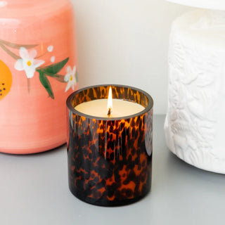 Candle in a tortoiseshell-patterned glass with a pink floral container and white textured object in the background.