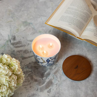 Candle in a decorative holder with a wooden lid, flowers, and an open book on a textured surface.